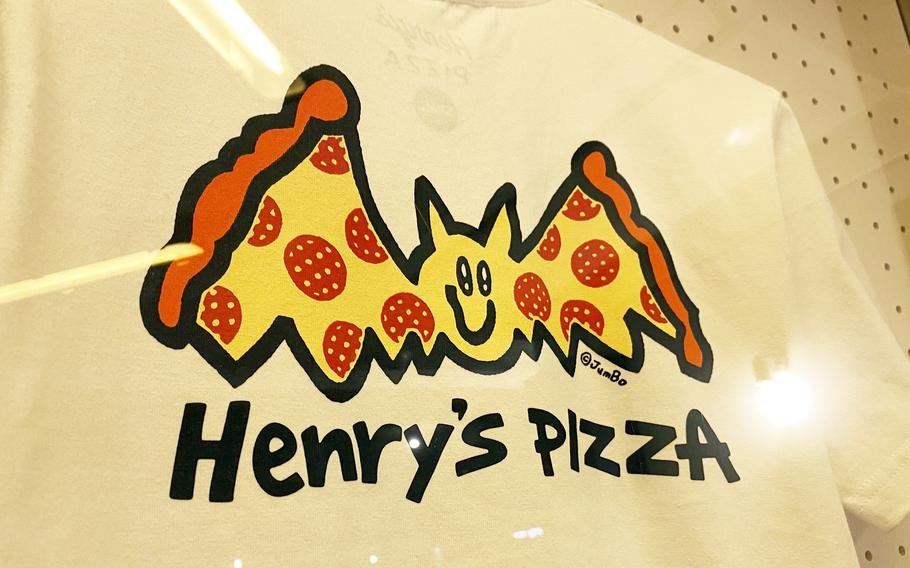 A close-up of a T-shirt logo shows a yellow bat with wings made out of pizza slices, and the stylized words ‘Henry’s Pizza’ below it.
