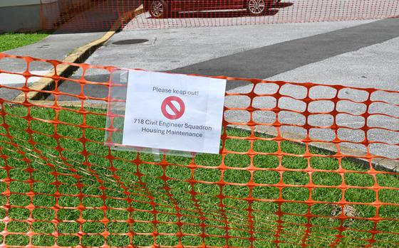 Orange safety fence with a white sign reading ‘Please keep out!’ and a red prohibition symbol, posted by 718 Civil Engineer Squadron Housing Maintenance. The fence blocks access to a grassy area near a sidewalk and street.