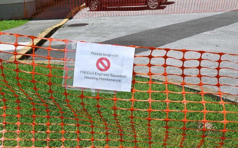 Orange safety fence with a white sign reading ‘Please keep out!’ and a red prohibition symbol, posted by 718 Civil Engineer Squadron Housing Maintenance. The fence blocks access to a grassy area near a sidewalk and street.