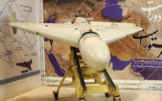 A military drone sits on a metal stand in a display in front of a wall with a map of desert terrain and Arabic writing.