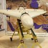 A military drone sits on a metal stand in a display in front of a wall with a map of desert terrain and Arabic writing.
