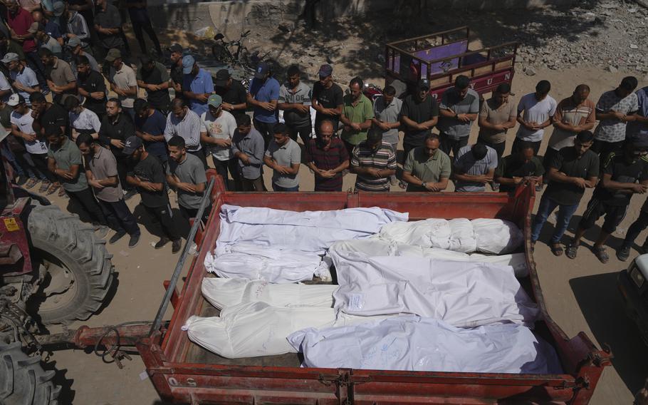An aerial view of rows of mourners praying next to a cart holding multiple shrouded bodies.