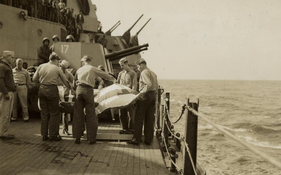 A black-and-white, 1940s-era image of sailors wrapping a body in a flag near the rail of a battleship as they perform a burial at sea.
