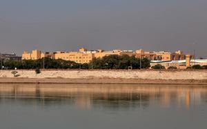 A government embassy building is seen from a distance with a river in the foreground and the skyline behind it.