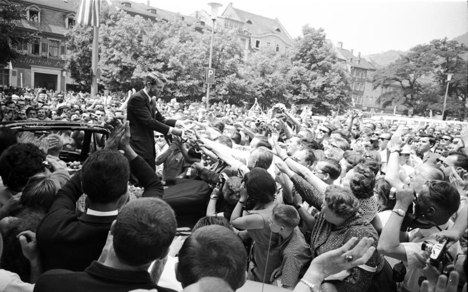 A black and white photo of a mass of people reaching their hands towards a Robert F. Kennedy. 