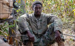A soldier in a camouflage uniform sits on the ground in a wooded area with his arms resting on his knees.