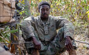 A soldier in a camouflage uniform sits on the ground in a wooded area with his arms resting on his knees.
