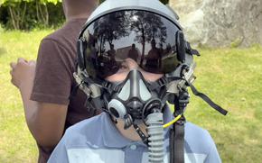 A young boy in a blue polo shirt looks at the camera while peeking out from beneath a fighter pilot helmet. 