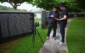 Two Japanese men in gray stand on a stone path and look down at a tablet next to a tripod in front of a dark marble monument engraved with lines of names in white.