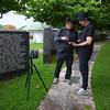 Two Japanese men in gray stand on a stone path and look down at a tablet next to a tripod in front of a dark marble monument engraved with lines of names in white.