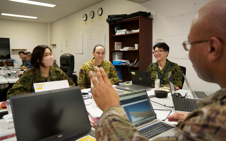 Military personnel in camouflage uniforms collaborate around a table with laptops and equipment in an office setting. 