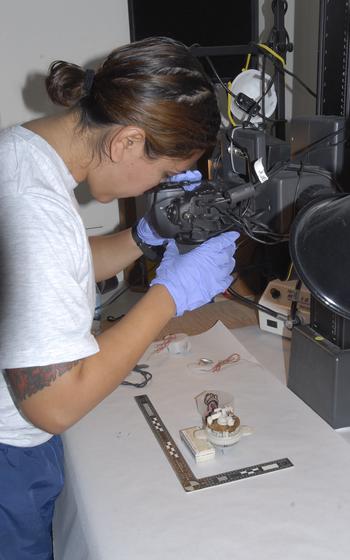 A vertical photo of a service member with medical gloves looking into a machine aimed at technical components on a white table.