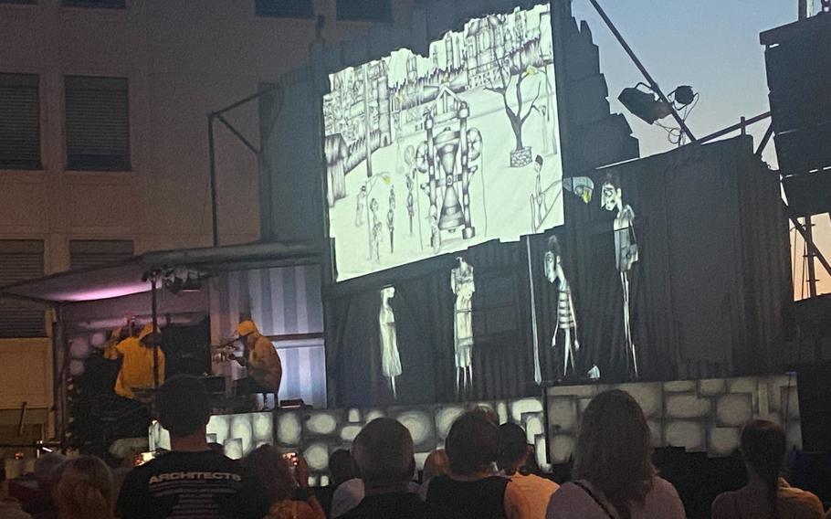 Standing members of an audience look up at a stage with a display and a band playing.