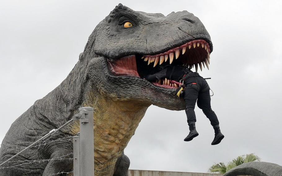 A person in black tactical gear dangles from the open mouth of a large, gray, animatronic Tyrannosaurus Rex, with prominent sharp teeth and yellow eyes against an overcast sky.