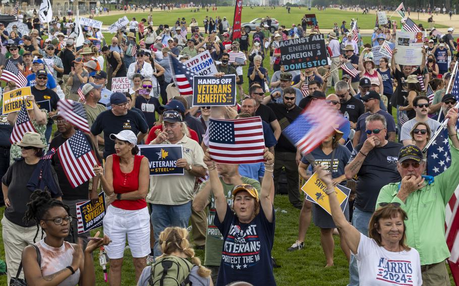 A large group of people, seen from above, stand on the National Mall with protest signs advocating for military veterans and waving American flags.