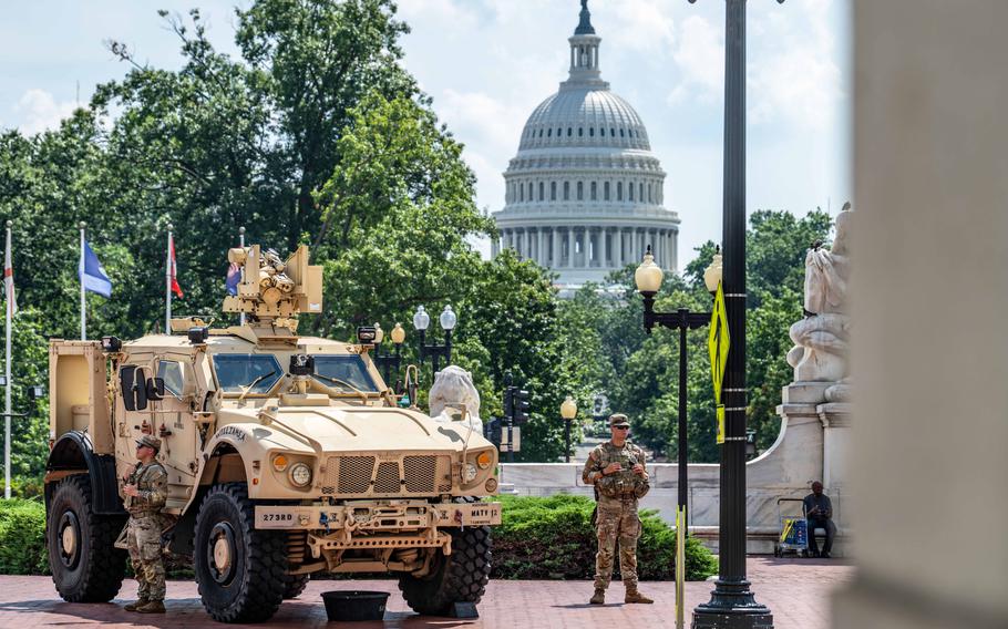 Two soldiers stand alongside a military vehicle with trees and the U.S. Capitol in the background.