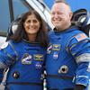 Suni Williams and Butch Wilmore stand outdoors wearing blue space suits covered with patches, including American flags and the NASA logo. 