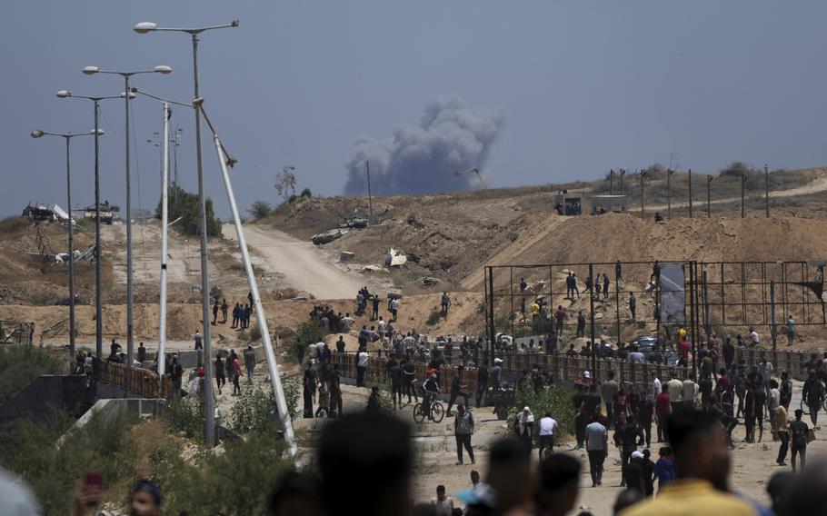 Dozens of Palestinians mill about in a humanitarian aid distribution center as smoke rises over a hill in the background.