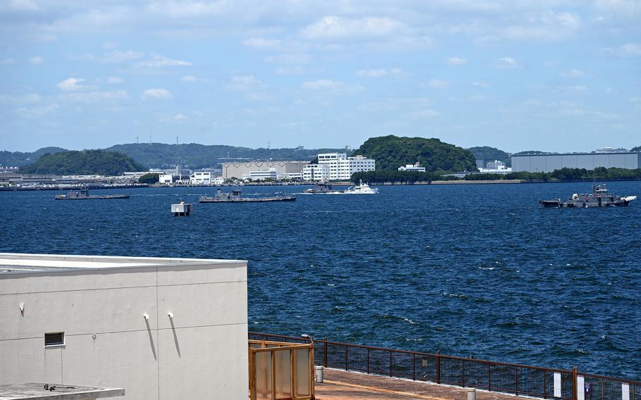 
A view showing a busy harbor with deep blue water. Multiple naval ships are visible in the distance, along with forested hills dotted with communication towers. Industrial and residential buildings line the shoreline, creating a typical naval base landscape.