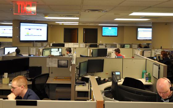 People working in cubicles in an office setting, with large monitors on the walls around them.