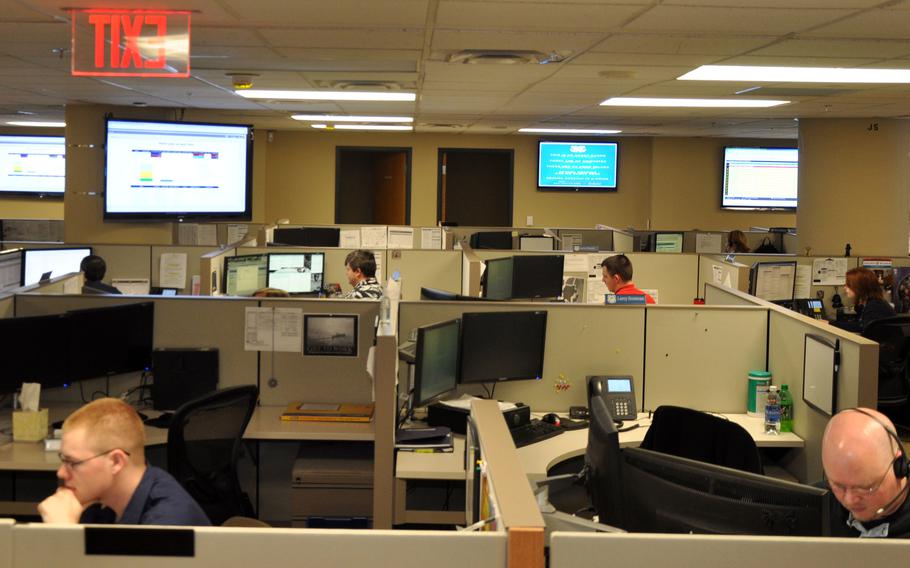 People working in cubicles in an office setting, with large monitors on the walls around them.