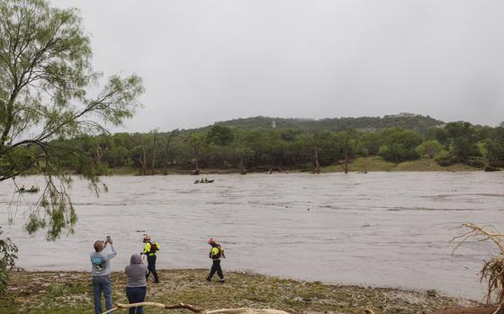 Rescue workers and volunteers stand on the shore of a river as boats travel in the water with a forested hill on the opposite side.