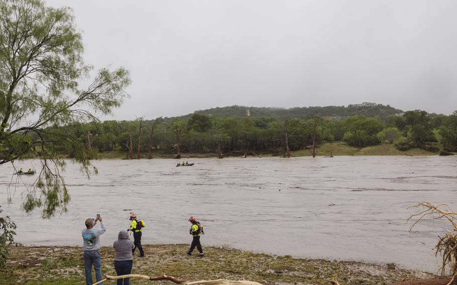 Rescue workers and volunteers stand on the shore of a river as boats travel in the water with a forested hill on the opposite side.