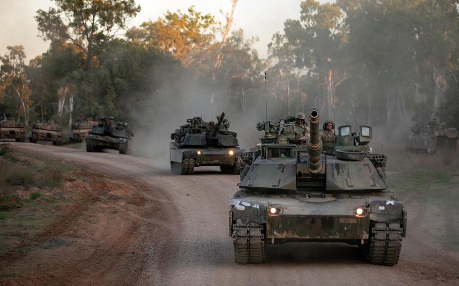 A convoy of military tanks moves along a dirt road, kicking up dust clouds. The lead tank has its headlights on and crew members visible in the turret. 