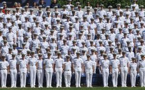 Rows of Naval Academy midshipmen in white dress uniforms and hats stand at attention during an outdoor graduation ceremony.