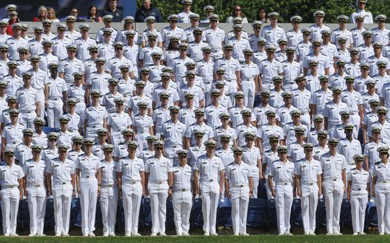 Rows of Naval Academy midshipmen in white dress uniforms and hats stand at attention during an outdoor graduation ceremony.