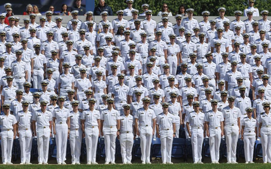 Rows of Naval Academy midshipmen in white dress uniforms and hats stand at attention during an outdoor graduation ceremony.