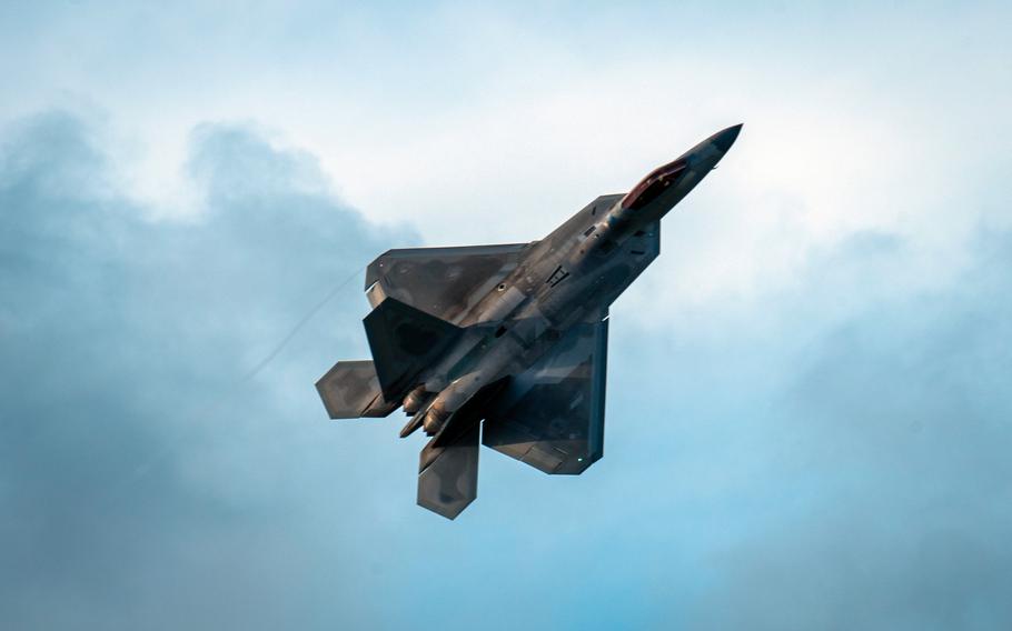 An F-22 Raptor flies directly overhead with a cloudy sky in the background.