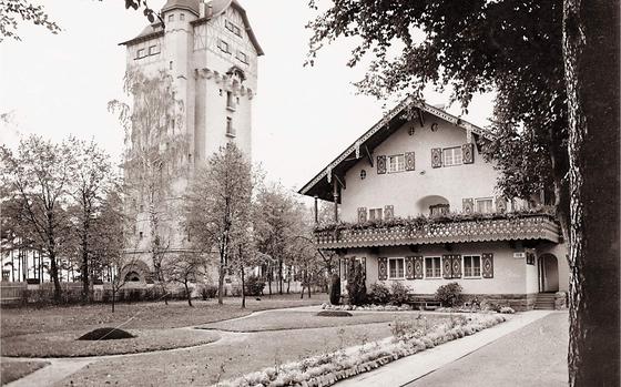 A black and white photo shows a tall brick building next to a nice home with a manicured lawn. 