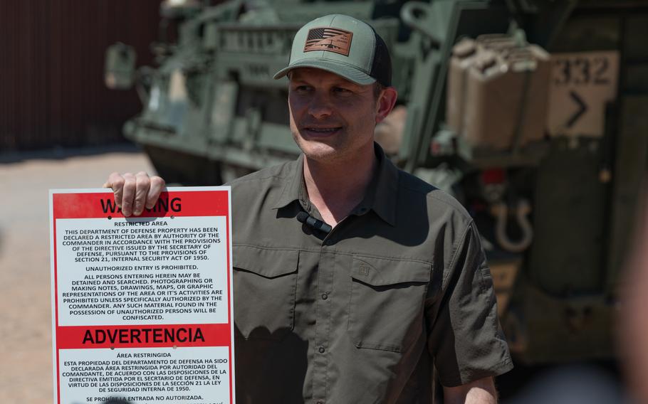 Hegseth holding up a sign with a military vehicle behind him. 