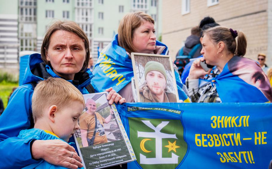 Three Ukrainian women hold photos of their missing relatives, while one hugs a boy at an outdoor event where Ukrainian soldiers arrived after a POW exchange with Russia. 