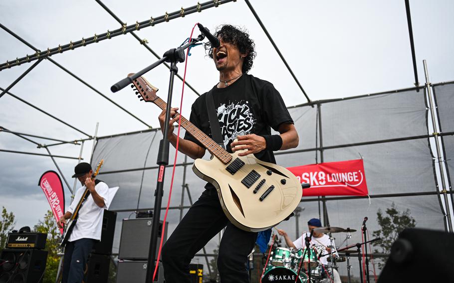 A Japanese man in a black T-shirt plays a guitar and sings into a microphone on a stage with a drummer and another guitarist behind him.