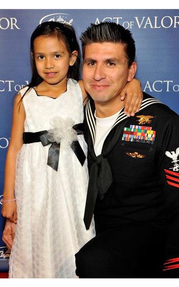A man in a Navy uniform with a young girl against a poster for the film “Act of Valor.”