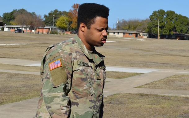 A man in a camouflage uniform walks across a grassy area with buildings and the treeline in the background.