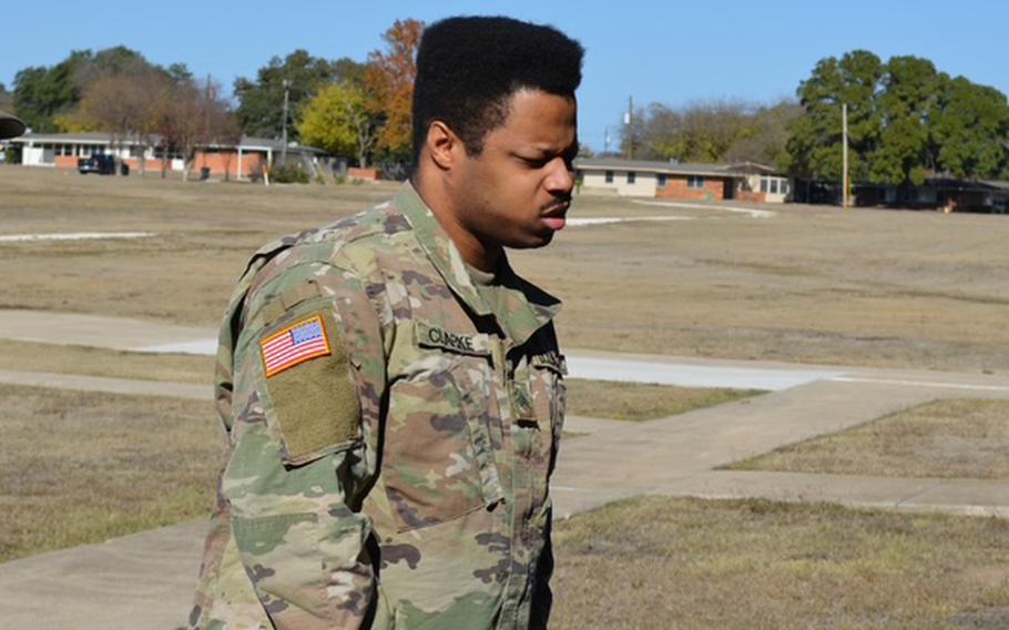 A man in a camouflage uniform walks across a grassy area with buildings and the treeline in the background.