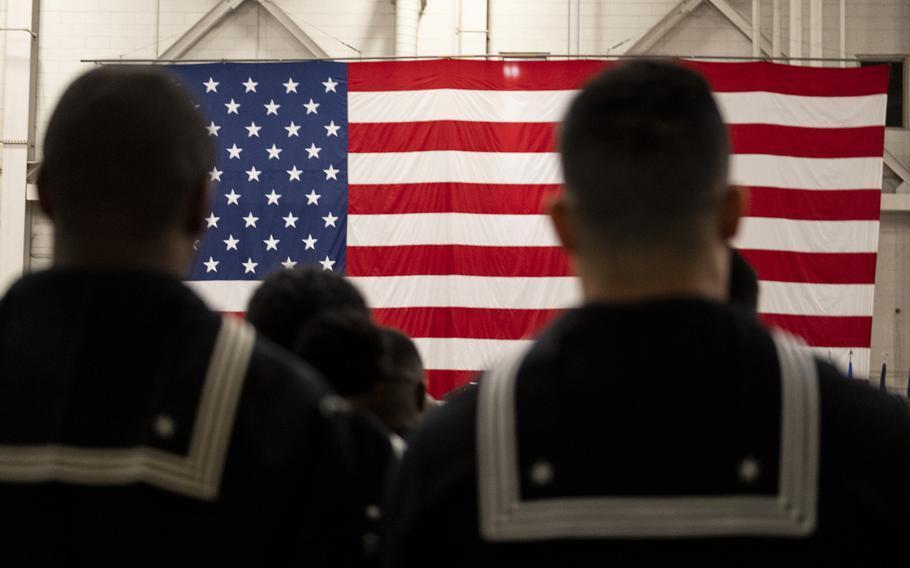 Sailors are seen from behind while they stand in front of an American flag.