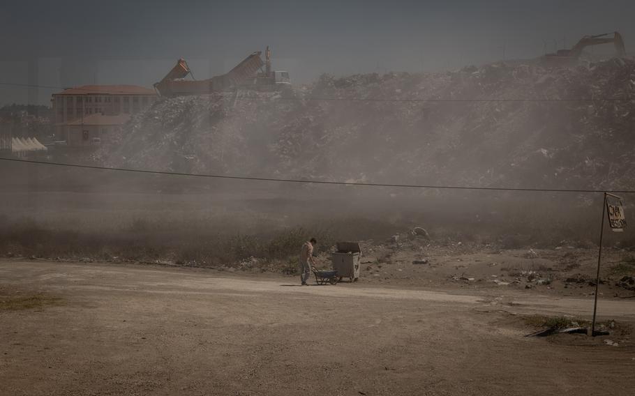 A restaurant worker in Samandag, Turkey, disposes of trash amid a cloud of dust from rubble being dumped on a site just a stone’s throw from the establishment. 