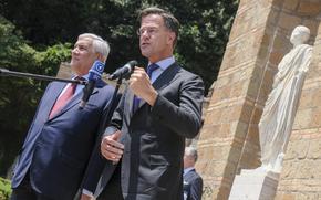 Two men in suits speak before microphones, in front of a statue.