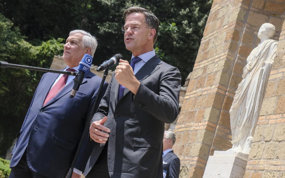 Two men in suits speak before microphones, in front of a statue.