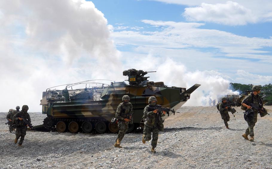 Troops in camouflage combat gear carrying rifles disperse from an armored vehicle on a beach, as smoke billows into the air in the background.