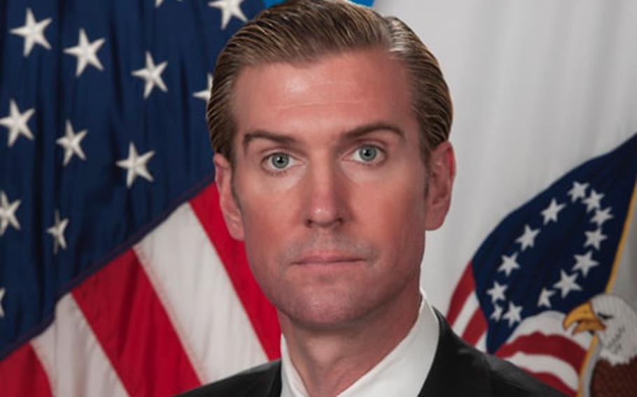 A man in a dark suit posing for a government portrait in front of a U.S. flag.