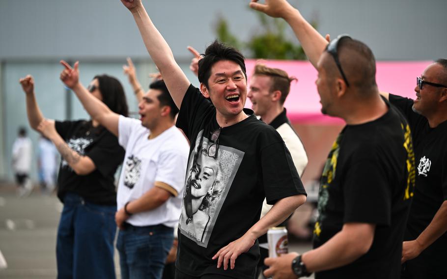 Spectators in black and white T-shirts raise their arms and cheer while standing in a crowd.