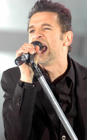 A man in a black suit and dress shirt sings into a microphone.