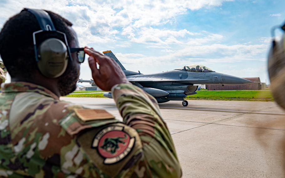 A military service member in camouflage uniform and headset salutes a fighter jet on an airport tarmac. 