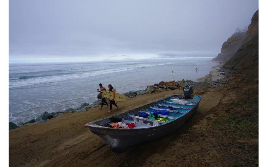 Surfers walk past one of two pangas that are believed to have been used to smuggle migrants and overturned in the ocean where at least eight people died.