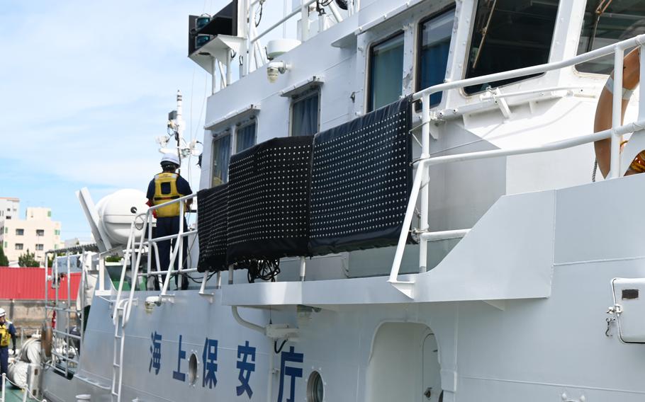 A white coast guard ship docked at port, showing the superstructure with Japanese characters visible on the hull. A crew member in a yellow safety vest is working on deck.